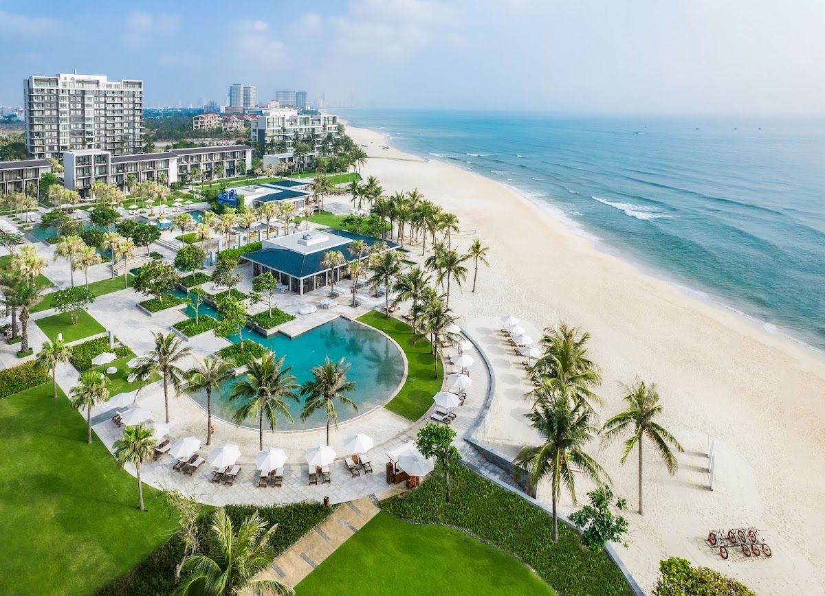 Aerial view of a luxury beachfront resort on Non Nuoc beach in Da Nang showing a curved pool, palm trees, sun loungers, and the white sand shoreline Aerial view of a luxury beachfront resort on Non Nuoc beach in Da Nang showing a curved pool, palm trees, sun loungers, and the white sand shoreline