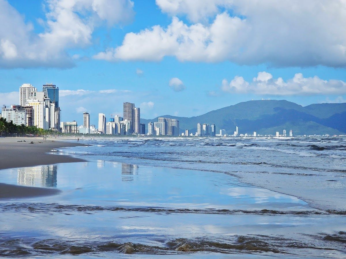 My Khe Beach with gentle surf and Da Nang's city skyline and green mountains reflected in the wet sand